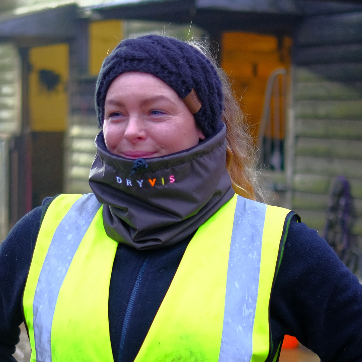 A woman outdoors wearing a brown DRYVIS fleece-lined waterproof snood, along with a black knit beanie and a high-visibility yellow safety vest.

