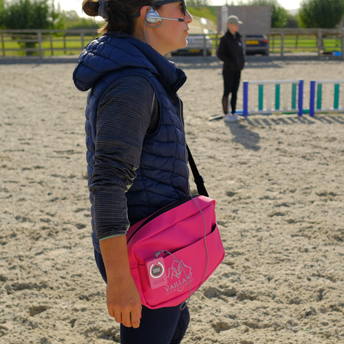 A female equestrian coach in a navy puffer vest and grey long-sleeve shirt stands in an outdoor sand arena wearing a bright pink Vaillant Equine Performance-embroidered crossbody bag made by DRYVIS. She is wearing a headset, and another person is visible in the background near horse jumps.
